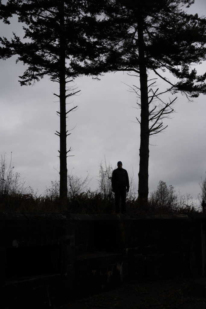 Dave standing between two tall evergreens, near the edge of a bluff at Fort Flagler, WA